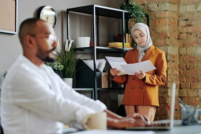 A woman wearing a hijab standing and reviewing documents in a modern office while a colleague works at his desk.